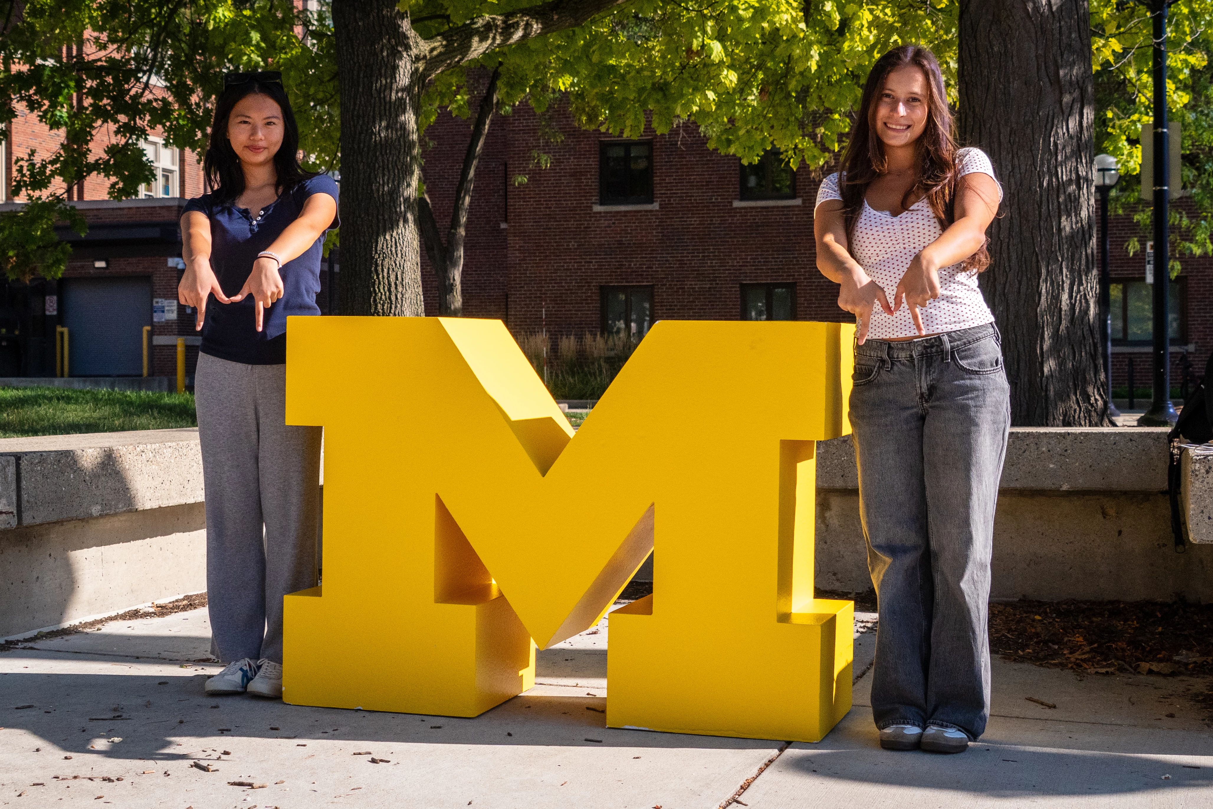 Two students standing next to a large wooden block M. The students are making M shapes with their fingers. 